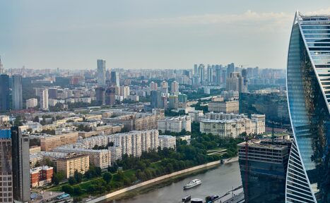 Арендовать Свидания в Москва Сити. Вид с высоты на городской пейзаж с реками и зданиями, в том числе многоэтажки и офисные небоскрёбы. На переднем плане река с яхтой, справа современное высотное здание с изогнутым фасадом. День, облачное небо.