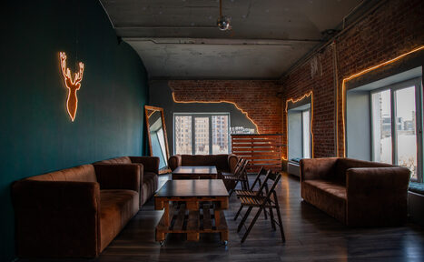 Арендовать ЛОСЬ - ламповый уютный лофт для небольшой компаний с видоя на Яузу. Лофт-пространство с кирпичными стенами, деревянным полом и диванами коричневого цвета. На стене неоновая вывеска в виде головы оленя. Разделочные столы и стулья из темного дерева. Освещение представлено неоновыми линиями и естественным светом из окон.