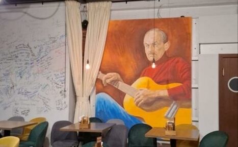 Арендовать Гнездо Глухаря. Кафе или пространство для встреч с деревянными столами и разноцветными стульями. На стене большое изображение человека с гитарой. Светлые занавески отделяют зону. Подвесные лампы создают мягкое освещение. На стене виден текст.