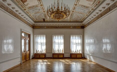 Арендовать Особняк Сухозанета. Просторная комната с паркетным полом, богато украшенным потолком с люстрой. Три высоких окна, через которые проникает свет, задрапированы полупрозрачными шторами. Стены светлые, украшенные декоративной лепниной.