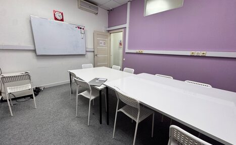 Арендовать Зелёный кабинет. Комната для встреч с белыми стенами и одной фиолетовой стеной, большое белое прямоугольное стол с белыми пластиковыми стульями, доска для маркеров, кондиционер, часы, розетки, серая ковровая плитка на полу.