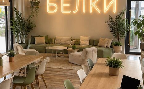 Арендовать БЕЛКИ. Просторное помещение с деревянными столами и стульями, зеленый диван с подушками, кресла, растения в горшках. На стене неоновая надпись. Большие окна, светлые стены, лампы и естественное освещение.