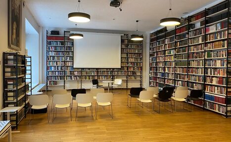 Арендовать Зал для вашего мероприятия. Помещение библиотеки с книжными стеллажами вдоль стен, проекторным экраном впереди. Расставлены стулья в несколько рядов. Потолочные светильники, светлый деревянный пол, белые стены. Видно большое окно слева.