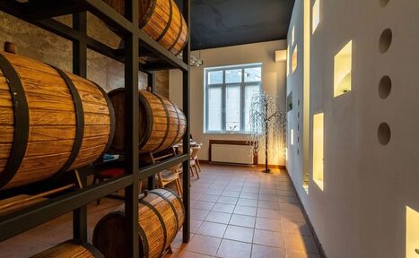 Арендовать La Cueva room. Пространство с деревянными бочками, стеллажами, столом и стульями. Терракотовая плитка на полу, белые стены с квадратными нишами, большое окно. Освещение встроенное и естественное. Потолок темного цвета.