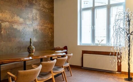 Арендовать La Cueva room. Просторная комната с деревянным столом и четырьмя стульями, стены с текстурой под бетон, большие окна с жалюзи, радиатор, декоративное дерево, пол из плитки коричневого цвета, ваза на столе.