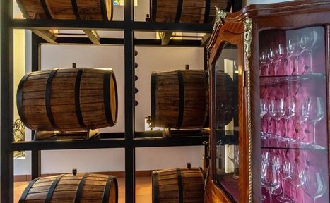 Арендовать La Cueva room. Деревянные бочки на металлических полках в винном погребе. Справа стеклянный шкаф с бокалами. Освещение мягкое, естественное. Цвета: дерево тёмных оттенков, металлические элементы, белые стены.