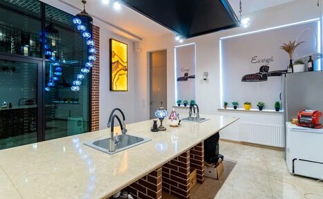 Арендовать La Cueva room. Кухонное пространство с длинным островом и двумя встроенными раковинами. Окружено кирпичными панелями и подвесными светильниками с синими элементами. На стене декор в виде панно и светодиодной ленты. Есть холодильник, декор и растения.