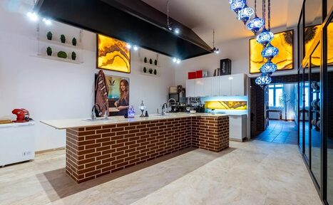 Арендовать La Cueva room. Кухонное пространство с островом, облицованным кирпичом. Белые шкафы, встроенные светильники, декоративные панели с подсветкой. Подвесные лампы синих оттенков. Плиточный пол, светлые стены. Видны кухонные приборы и декор.