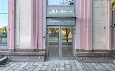 Арендовать QuintesSense. Вход в здание с двойными стеклянными дверями, серыми рамами. Над дверями вывеска с двумя текстовыми строками, верхняя содержит неразборчивый текст, нижняя — "Центр Ресурсного Состояния". Стены светло-серого цвета с розовыми колоннами.