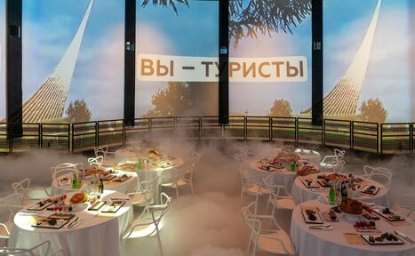 Арендовать Атракцион для различных событий. Круглое помещение с проекцией на стенах, изображающей шпили и текст "Вы - туристы". В центре столы с белыми скатертями, сервированными для банкета. Белые стулья, искусственный туман на полу.