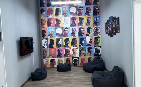 Арендовать Лофт Радуга. Комната с серыми стенами, одна из которых украшена яркими постерами с изображениями персонажей. На полу деревянное покрытие, имеются пять мягких кресел-мешков, на одной стене телевизор. Освещение потолочное, на стене постер с изображениями.