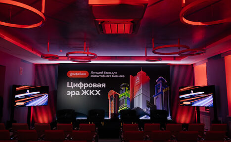 Арендовать Дом Жилищного Работника. Конференц-зал с рядами кресел, двумя экранами слева и справа, цветной баннер на заднем плане с городской сценой. Красное и фиолетовое освещение, потолочные кольцевые светильники. Стены с вертикальными панелями.