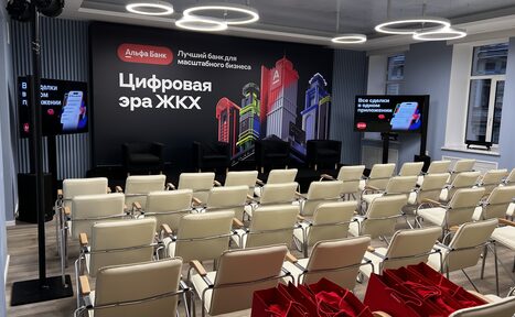 Арендовать Дом Жилищного Работника. Аудиторное пространство с рядами белых стульев, черной сценой с креслами, задником с текстом и изображениями зданий. На стенах светодиодные экраны, один с синим интерфейсом. Круглые светильники на потолке, серые стены в полоску. Красные пакеты на полу.