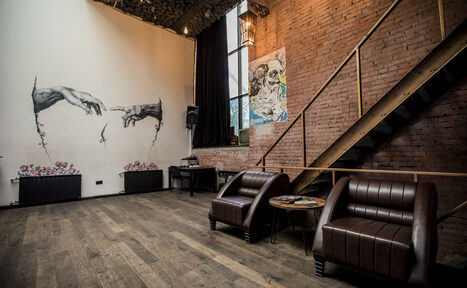 Арендовать Street Art Loft (Стрит Арт Лофт). Лофт-пространство с кирпичными стенами и деревянным полом. Два кожаных кресла и круглый столик. На одной стене граффити, на другой — картина. Металлическая лестница и большие окна с тяжелыми шторами. Высокие потолки, точечное освещение.