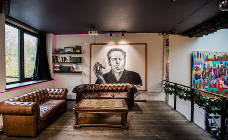 Арендовать Street Art Loft (Стрит Арт Лофт). Лофт-пространство с кожаными диванами и деревянным столиком на фоне портрета на стене, книжные полки, большое окно, яркая картина справа, пол из тёмного дерева, чёрный потолок, проектор, светильники, зелёный декор на перилах.