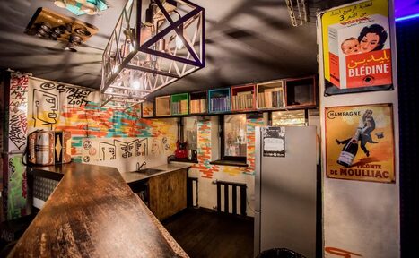 Арендовать Street Art Loft (Стрит Арт Лофт). Барное пространство с деревянной стойкой и стеллажами. Стены украшены граффити и постерами с надписями. Плиточные стены бежевого и разноцветного цвета. Металлические элементы освещения. Окна с черными рамами. Холодильник серого цвета.