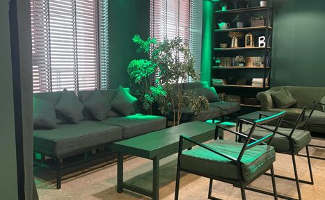 Арендовать Work Shop Loft(Воркшоп Лофт). Зал 11 . Современная гостиная в зелёных тонах с диванами и креслами, кофейным столиком, стеллажом с декором. На полу светлая поверхность, стены темно-зелёные, окна с жалюзи, комнатные растения добавляют уюта.