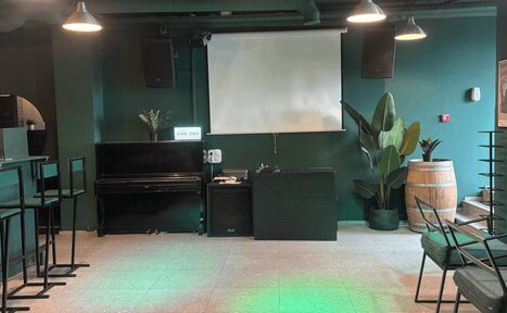 Арендовать Work Shop Loft(Воркшоп Лофт). Зал 11 . Бар с темно-зелеными стенами, столами и стульями в индустриальном стиле. На стене развешаны два громкоговорителя и большой экран. Справа - деревянная бочка и растения. Потолочные светильники создают мягкое освещение. На полу плитка.