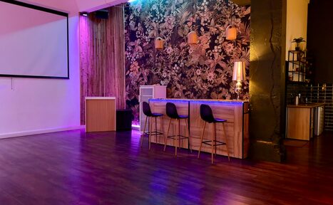 Арендовать Лофт для вечеринок с коктейльным баром . Большое помещение с деревянным полом и цветной подсветкой. Справа барная стойка, три барных стула, декоративные светильники и лампа. На задней стене цветочные обои. Слева настенный экран. Декоративная деревянная панель.