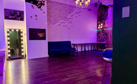 Арендовать Лофт для вечеринок с коктейльным баром . Просторная комната с деревянным полом и высоким потолком. Справа барный стул, слева диван. На стене светящаяся картина и декор из кирпича. Зеркало с подсветкой, люстра. Около стены длинные столы. Фиолетовая атмосфера освещения.