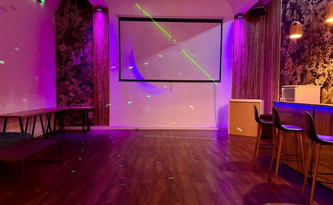 Арендовать Лофт для вечеринок с коктейльным баром . Просторное помещение с паркетным полом, столами и стульями, оборудовано проектором и экраном. Стены украшены темными цветочными обоями. Фиолетовое и розовое освещение. Видны деревянные панели и высокие стулья у барной стойки.
