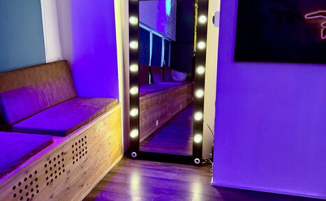 Арендовать Лофт для вечеринок с коктейльным баром . Компактное пространство с деревянным полом, на стене зеркало с лампочками, фиолетовое и синее освещение. Слева деревянная скамья с подушками, на стене справа абстрактное искусство.