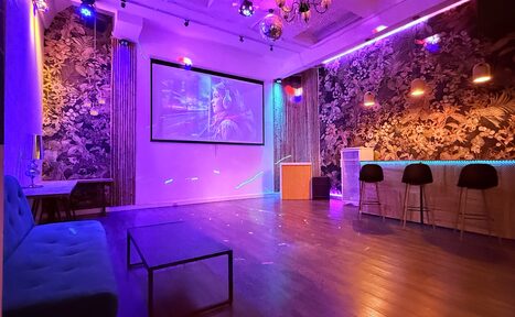 Арендовать Barshowplace (БарШоуПлейс). Лофт для вечеринок с коктейльным баром . Просторная комната для мероприятий: проектор с экраном на стене, барная стойка с тремя стульями, мягкий диван, низкий столик. Стены с цветочным узором, разноцветное освещение. Потолок с подвесными светильниками и диско-шаром. Пол из тёмного дерева.