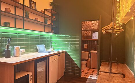 Арендовать Barshowplace (БарШоуПлейс). Лофт для вечеринок с коктейльным баром . Кухонное пространство с деревянной столешницей, мойкой и посудомоечной машиной. Зелёная плитка на стене с подсветкой. Полка с декором. Открытая вешалка с вешалками. Видна часть туалета с плиточным полом. Тёплое освещение.