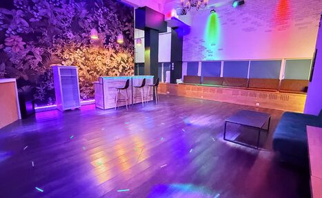 Арендовать Barshowplace (БарШоуПлейс). Лофт для вечеринок с коктейльным баром . Просторное помещение с деревянным полом, барной стойкой и высокими стульями, диванами вдоль стены. Освещение разноцветное, с акцентом на стенах. Темные обои с растительным узором. Потолок украшен люстрой.