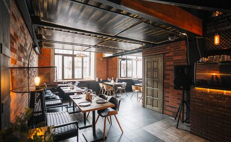 Арендовать Двухуровневый лофт-бар. Пространство для кафе с кирпичными стенами и металлическим потолком. Расставлены деревянные столы и смешанные стулья из дерева и кожи. Большие окна обеспечивают дневное освещение. Потолочные лампы и торшеры добавляют искусственный свет.