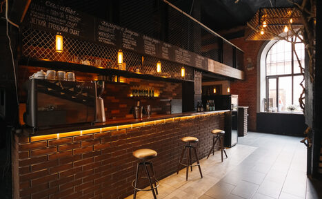 Арендовать Двухуровневый лофт-бар. Барная зона с кирпичной стойкой и тремя табуретами, черные сетчатые перегородки, коричневая плитка, подвесные светильники с теплым светом, большая арка с окнами, меню написано мелом на досках, металлические элементы интерьера, стены из красного кирпича.