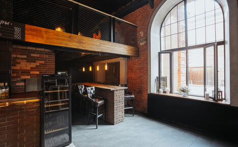 Арендовать Двухуровневый лофт-бар. Лофт пространство со столиком барного типа и высокими стульями, кирпичные стены, большой арочный оконный проем, светильники подвесного типа, пол серого цвета, барная стойка обложена плиткой, металлические и деревянные элементы в отделке.