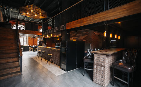 Арендовать Двухуровневый лофт-бар. Пространство в стиле лофт с кирпичными стенами и темными деревянными элементами. Два уровня с лестницей. Барная стойка с высокими стульями. Подвесное освещение, пол из серой плитки, черный потолок. Столы и стулья в зоне отдыха.