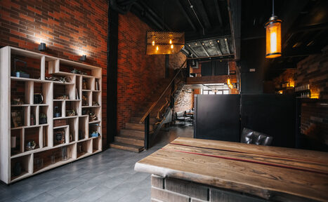 Арендовать Двухуровневый лофт-бар. Лофт-пространство с кирпичными стенами и деревянной меблировкой. Видна лестница, ведущая на второй уровень, деревянный стол перед стеллажом с полками, кресло. Освещение подвесными лампами, теплый свет. Пол из серой плитки.