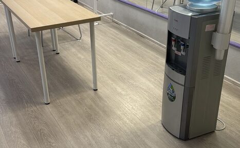 Арендовать Конференц-зал до 90 гостей. Пространство с деревянным столом и кулером для воды. Светлое напольное покрытие, серые стены, зеркало вдоль стены. Освещение естественное, мебель минималистичная. Электрическая розетка и провода на стене.