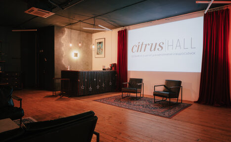 Арендовать Citrus Hall (Цитрус Зал). Grapefruit (Грейпфрут) . Просторное помещение с деревянным полом, черным потолком и красными занавесками. На стене крупный экран с надписью. Две черные кожаные кресла на ковре. Справа барная стойка. Освещение от подвесных ламп и светильников на стене.
