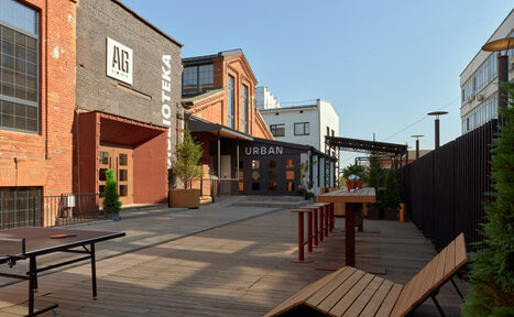 Арендовать AG Loft. Urban (АГ Лофт. Урбан). Просторный лофт с раритетным автомобилем. Площадка на открытом воздухе с деревянным настилом, длинными деревянными скамейками и столами, стол для пинг-понга. Здание из красного кирпича с большими окнами. Вывески "AG" и "URBAN". Планировка открытая, высокие стены. Растения в горшках.