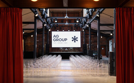 Арендовать AG Loft. Urban (АГ Лофт. Урбан). Просторный лофт с раритетным автомобилем. Большое помещение с высокими потолками, обставленное прозрачными стульями, расположенными рядами. На сцене установлен экран с надписью «AG GROUP». Стальные балки и прожекторы на потолке. Стены кирпичные, свет от подвесных ламп. Шторы красные.