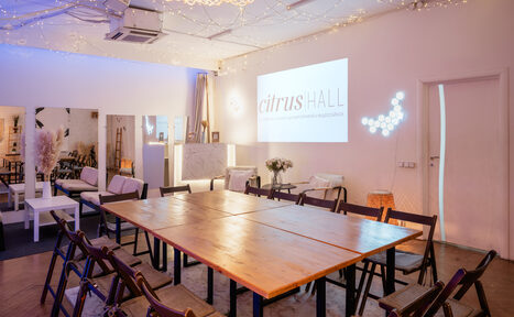 Арендовать Citrus Hall (Цитрус Зал). Sweetie (Конфетка). Пространство с деревянным столом в центре, окружённым складными стульями. На стенах светлые панели и проектор с изображением. В интерьере диваны, столики, декор, светодиодное освещение. Потолок с гирляндами. Тёплые тона, паркет.
