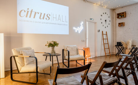 Арендовать Citrus Hall (Цитрус Холл). Зал Свити. Просторное светлое помещение с паркетным полом и белыми кирпичными стенами. Ряды деревянных стульев, кресла с подушками, стеклянный столик с вазой, настенные часы, лестница, проектор с изображением. Потолочные гирлянды и боковое освещение.