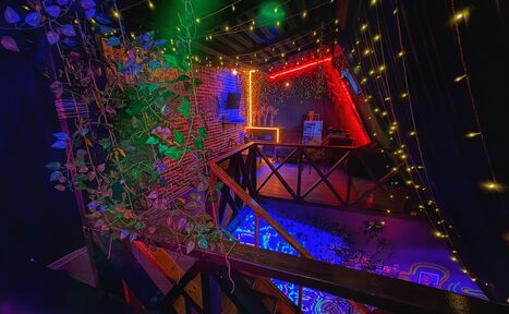 Арендовать Лофт-бар. Лофт-пространство с кирпичными стенами, деревянными перилами и балконом. Потолок украшен гирляндами. Зона освещена разноцветными неоновыми лампами. На мебельной полке несколько предметов, на стене плоский экран. Растения свисают с перил.