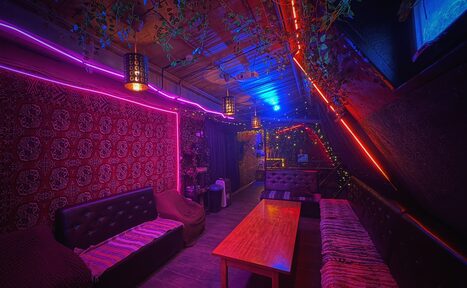 Арендовать Лофт-бар. Лофт с мягкой мебелью, включая диваны и кресла, деревянный стол. Стены с узорчатым ковром, освещение неоновыми и гирляндными огнями, разноцветные светильники на потолке. Потолочные балки декорированы растениями. Темные тона, яркие цветные акценты.