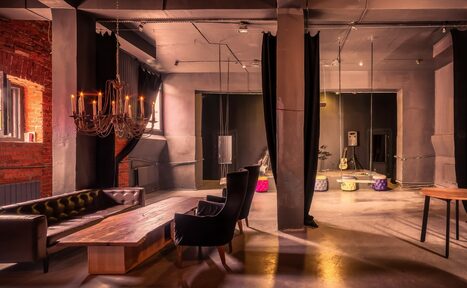 Арендовать Music loft (Мьюзик лофт). Лофт-пространство с открытой планировкой. Деревянный стол, кожаный диван, кресла, черные шторы. Лофт-освещение в виде люстры. Кирпичные и бетонные стены, зеркало, деревянный и бетонный пол. Цвета: коричневый, серый, черный.