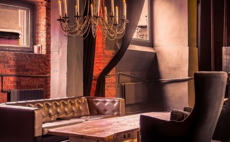 Арендовать Music loft (Мьюзик лофт). Просторное помещение с высокими кирпичными стенами. Коричневая кожаная диванная зона и деревянный стол. Большое окно с занавесью. Подвесная люстра с декоративными лампами. Теплое освещение. Элементы из металла и дерева, спокойные тона.