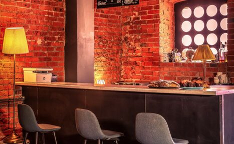 Арендовать Music loft (Мьюзик лофт). Барное пространство с кирпичными стенами, тремя серыми стульями у стойки и лампами с абажурами. На стойке корзинка с хлебом и предметы декора. Окно с круглым узором, мягкое освещение. Потемневшая труба в стене.