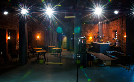 Арендовать Music loft (Мьюзик лофт). Лофт-пространство с кирпичными стенами, высокими потолками и промышленным освещением. В центре стойка с микрофоном. Есть диваны, круглые столы и барная стойка. Освещение яркое, зонирующее. Потолок из металлических конструкций. Цвета темные с акцентами.