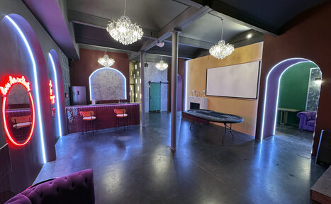 Арендовать Белый лофт с яркой мебелью. Просторное помещение с черным потолком и полом, украшенное сверкающими люстрами. В центре расположен длинный стол. Справа - белый экран для проектора. Ниша с барной стойкой и стульями. На стенах неоновые вывески, круглое зеркало с подсветкой.