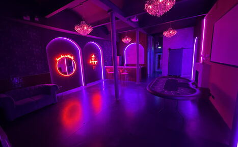 Арендовать Белый лофт с яркой мебелью. Современное пространство с неоновым освещением, тремя арками на стене, диваном слева, барной стойкой и табуретами посередине, покерным столом справа, люстрами на потолке. На стенах неоновые знаки. Цвета: фиолетовый, розовый.
