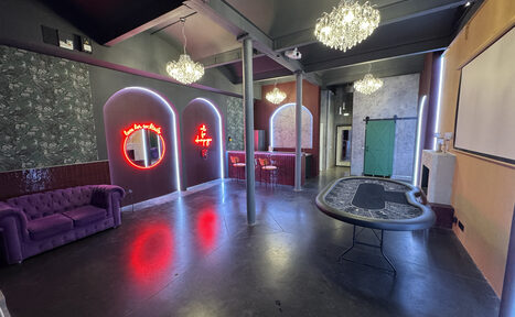 Арендовать Белый лофт с яркой мебелью. Просторная комната с высоким потолком, стол для игры, фиолетовый диван, барная стойка с табуретами, неоновая подсветка, декоративные арки, цветочные обои, зелёная раздвижная дверь, большие люстры. Пол тёмный, стены разноцветные.