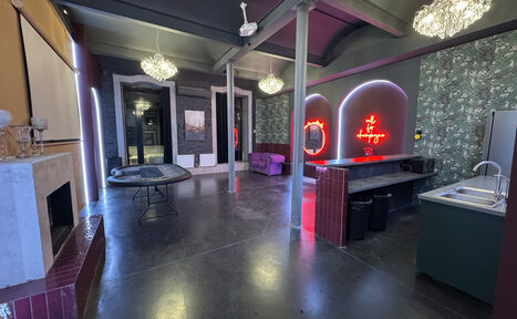 Арендовать Белый лофт с яркой мебелью. Просторное помещение с высокими потолками, бетонный пол. Барная стойка с раковиной, металлические колонны, камин, пуф, обеденный стол. Неоновый знак на стене, обои с узором. Люстры, темный потолок, большие окна с занавесками.